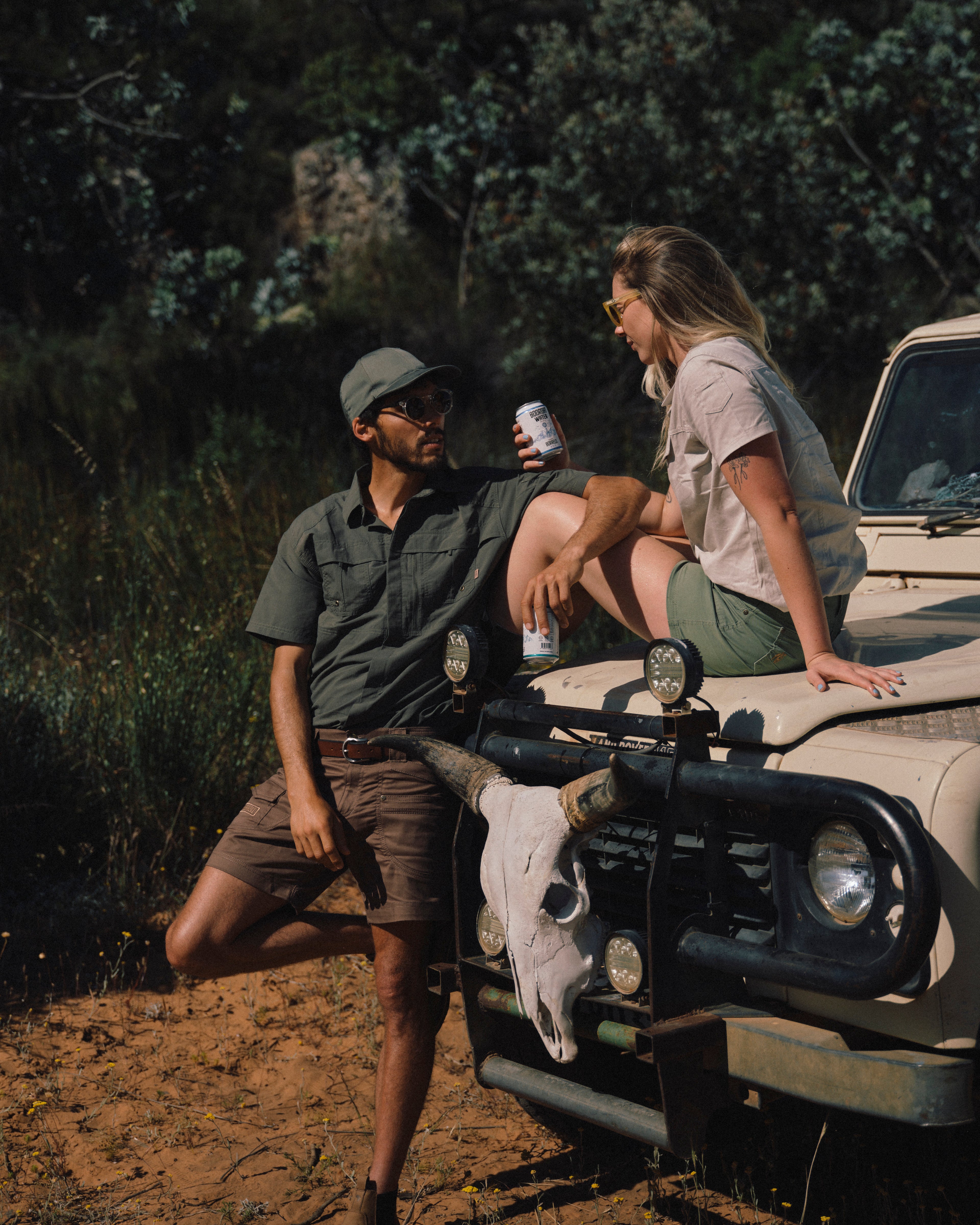 Two people sitting on a jeep with a skull decoration, surrounded by nature.